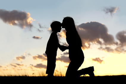 Silhouette of a young mother lovingly kissing her little child on the forehead, outside in front of a sunset in the sky.
